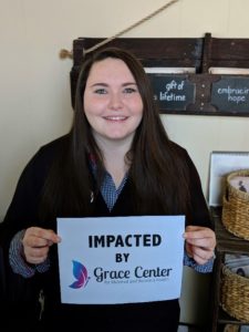 Young mother with brown hair smiling and holding a sign that reads Impacted by Grace Center
